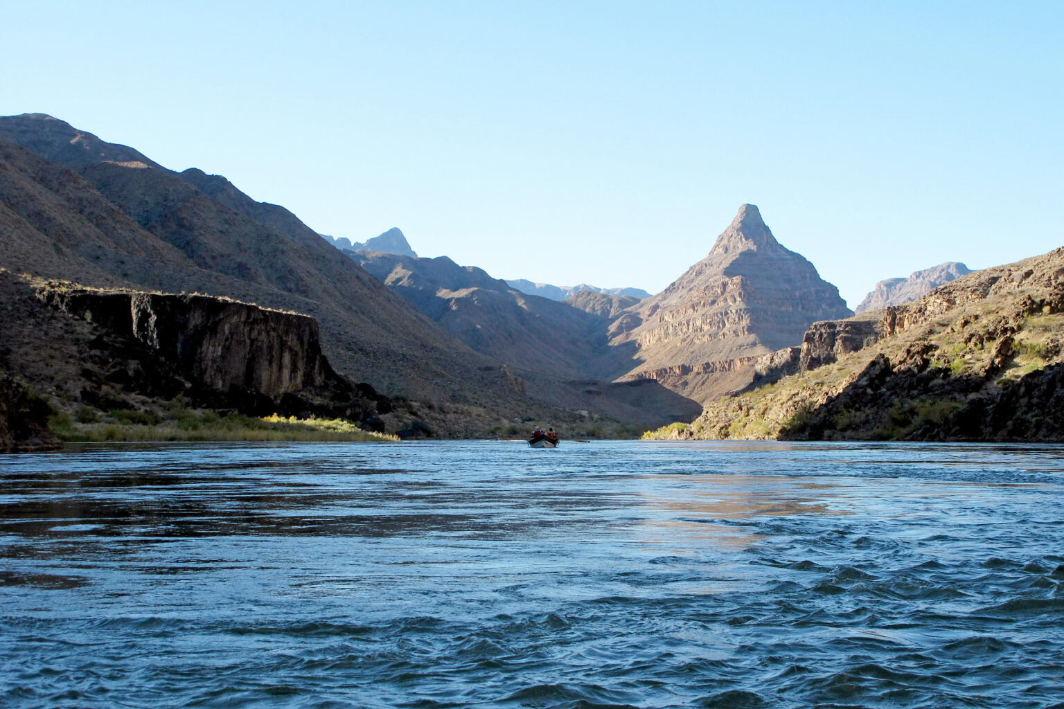 A lone dory floats down a wide, calm Colorado River in the lower section of Grand Canyon with a pyramid shaped mountain in the background