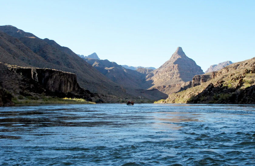 A lone dory floats down a wide, calm Colorado River in the lower section of Grand Canyon with a pyramid shaped mountain in the background