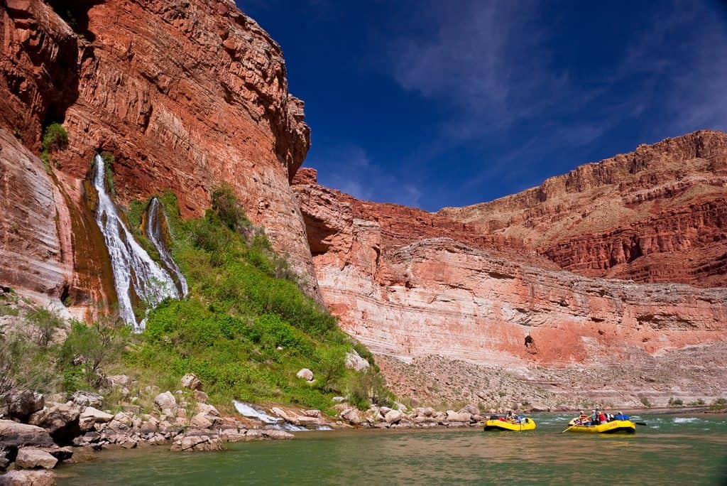 Two yellow rafts float down the Colorado River with Vasey's Paradise, a natural spring in Grand Canyon, gushing water off the cliffside